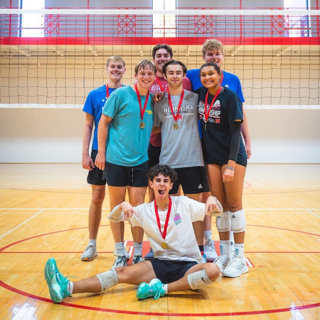 Seven Huskers wearing medals pose on the volleyball court.