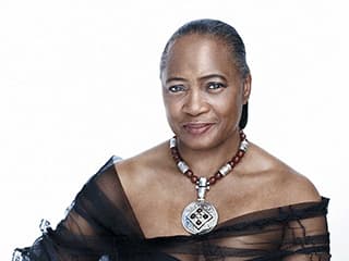 Portrait of Barbara Hendricks in a black dress and necklace against a solid, white backgound.